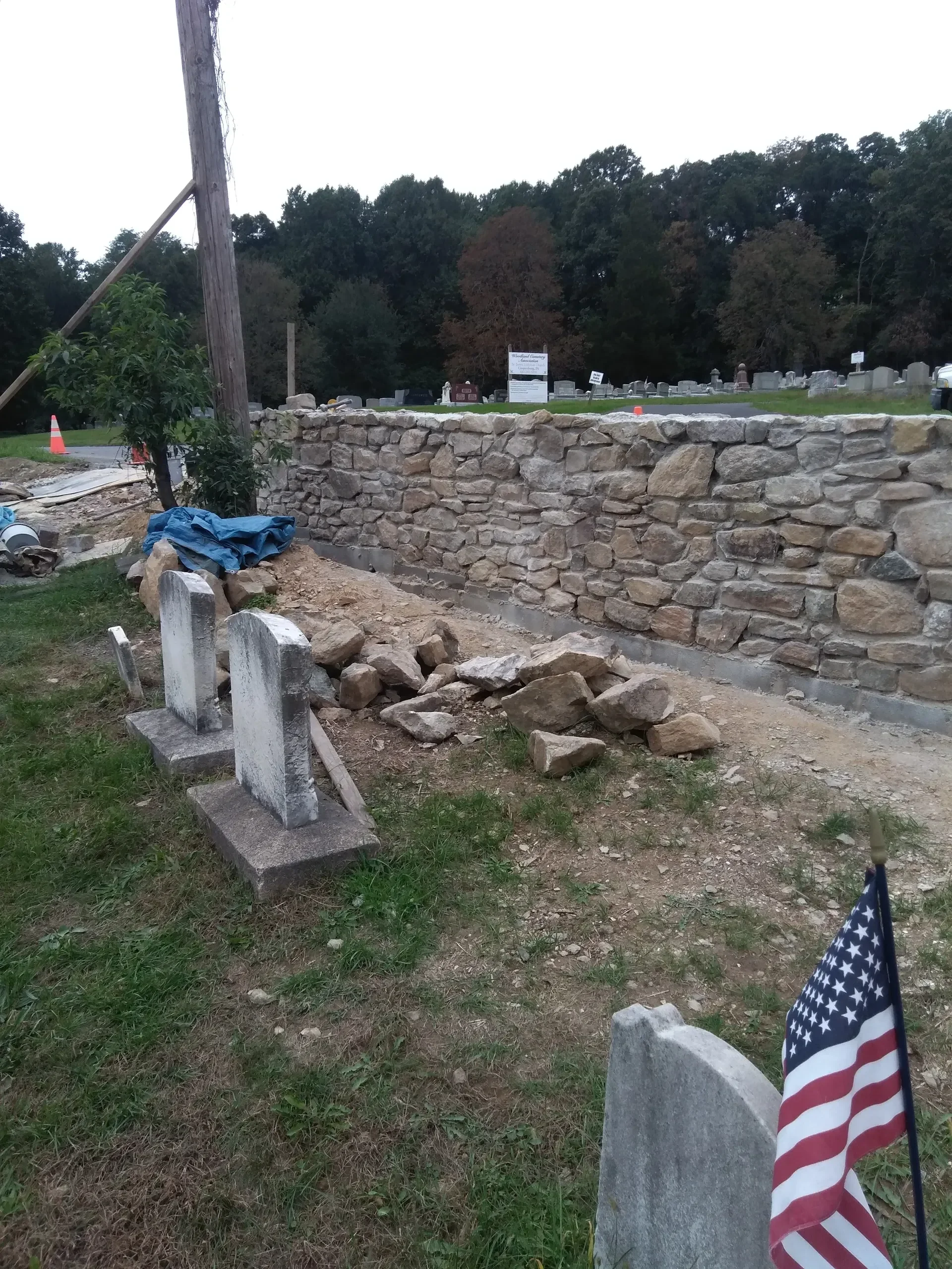 A newly constructed stone wall stands behind old gravestones amidst construction debris, indicating ongoing restoration work in the cemetery.