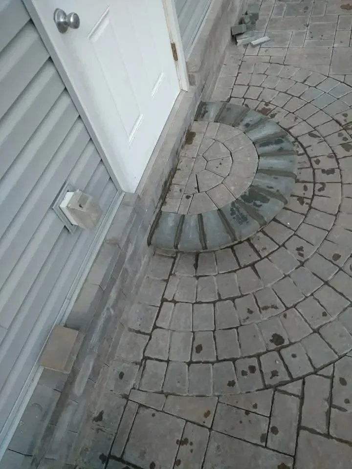 A close-up view captures a gray cobblestone paver patio entrance featuring a semi-circular step leading up to a white exterior door and light gray vinyl siding.