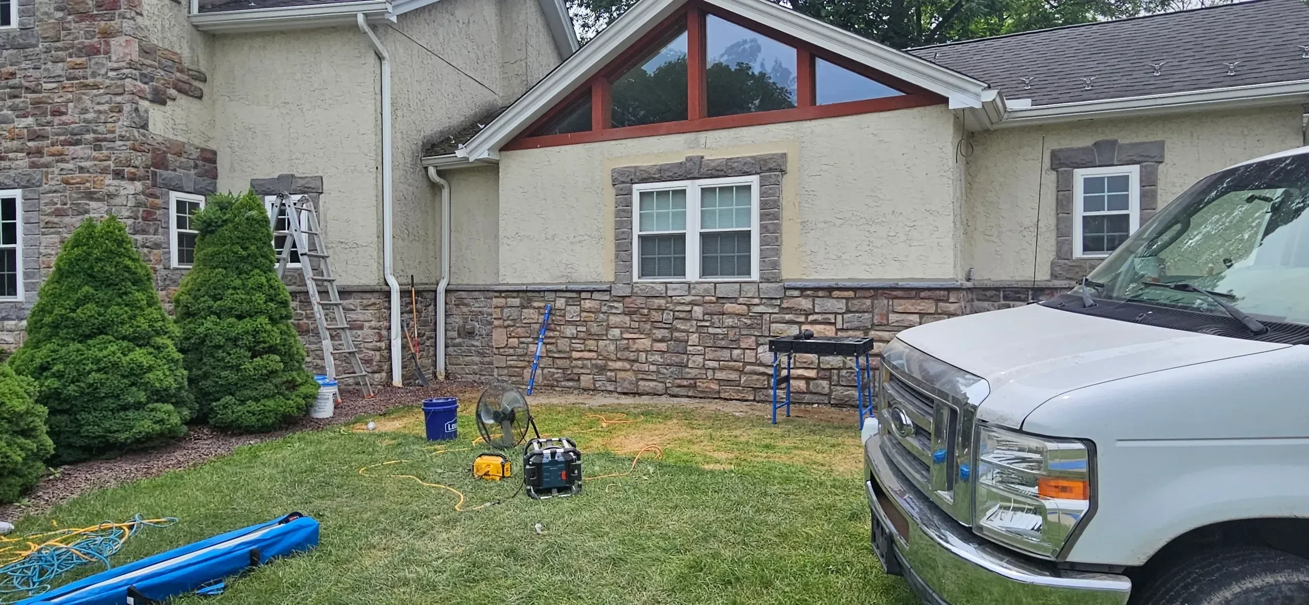 Construction equipment, including a ladder and power tools, sits on the lawn next to a house featuring stone veneer and stucco siding under renovation.
