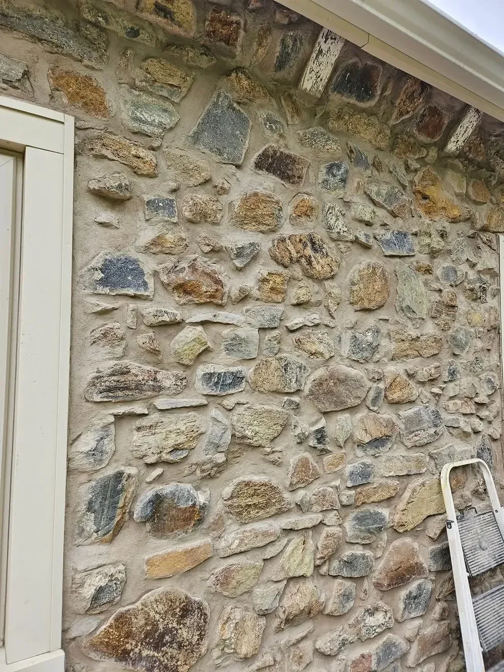 A textured exterior wall is covered in irregular, multicolored natural stones set in thick mortar, featuring a white window trim and a small step ladder leaning against the surface.