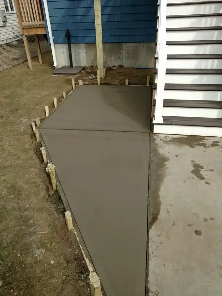 A newly poured, trapezoidal concrete slab is visible in a residential backyard, bordered by wooden stakes and adjacent to a house foundation and exterior stairs.