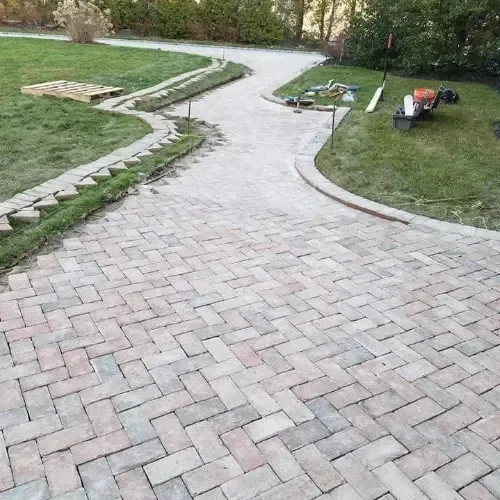A newly installed herringbone paver walkway curves through a residential lawn, with construction tools and unfinished edging visible along the grassy borders.