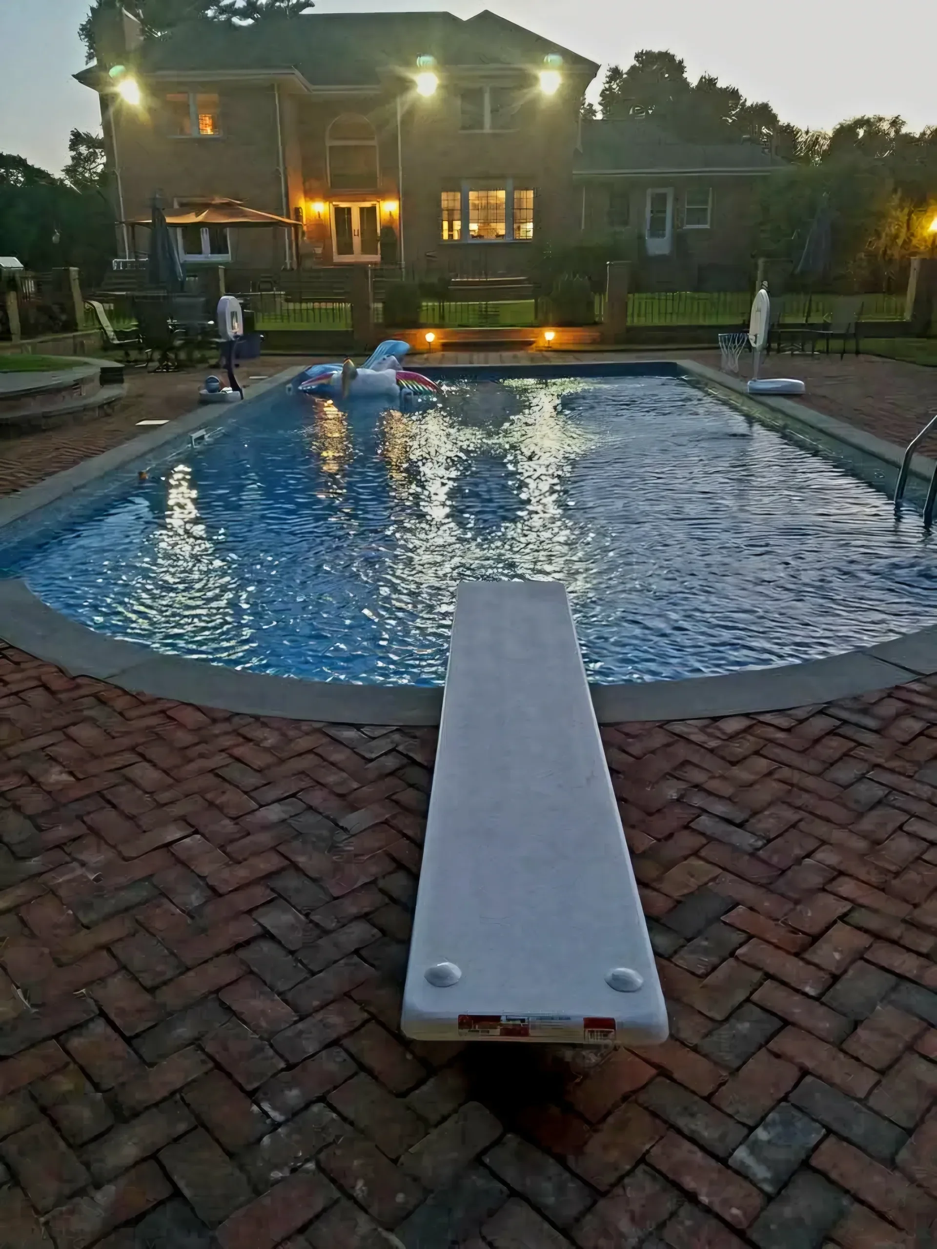 A residential swimming pool with a diving board and brick patio is brightly illuminated at dusk, featuring pool floats and a large house in the background.
