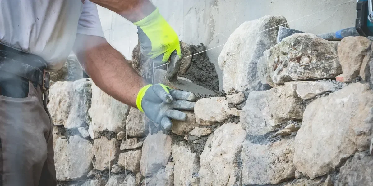 A mason applies mortar between irregular stones while building a rustic wall.