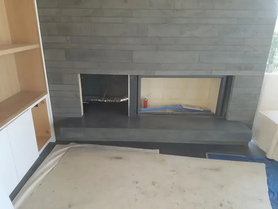 A modern fireplace clad in gray stacked stone is being installed next to light wood built-in cabinetry, with visible construction elements indicating the project is ongoing.