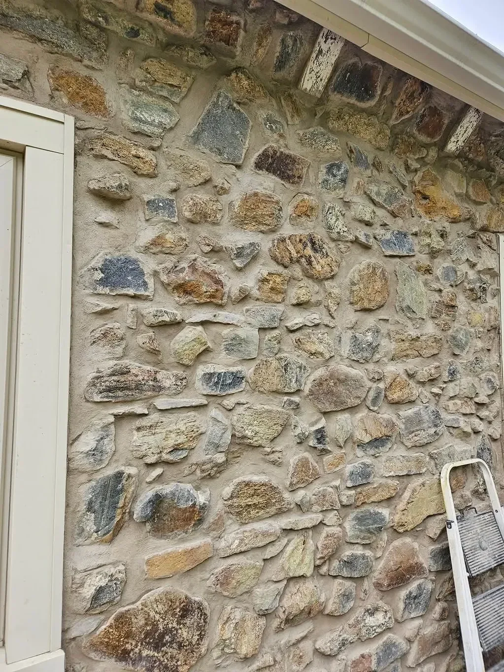 A detailed view of a rustic exterior wall features irregular, multi-colored natural stones set in thick mortar, with a white step ladder leaning against the finished surface.
