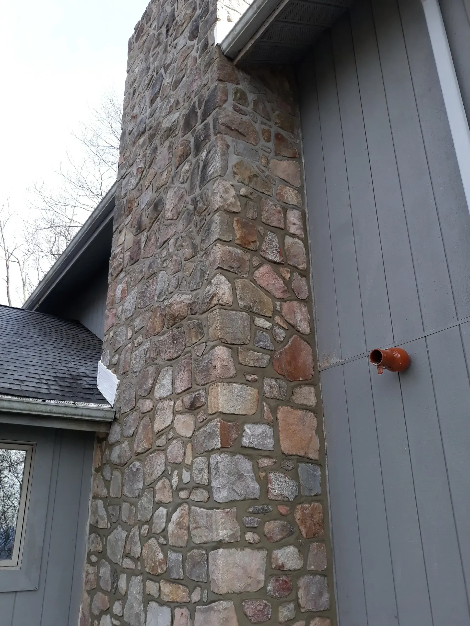 A tall residential chimney clad in multi-colored natural stone veneer stands next to vertical grey siding under an overcast sky.