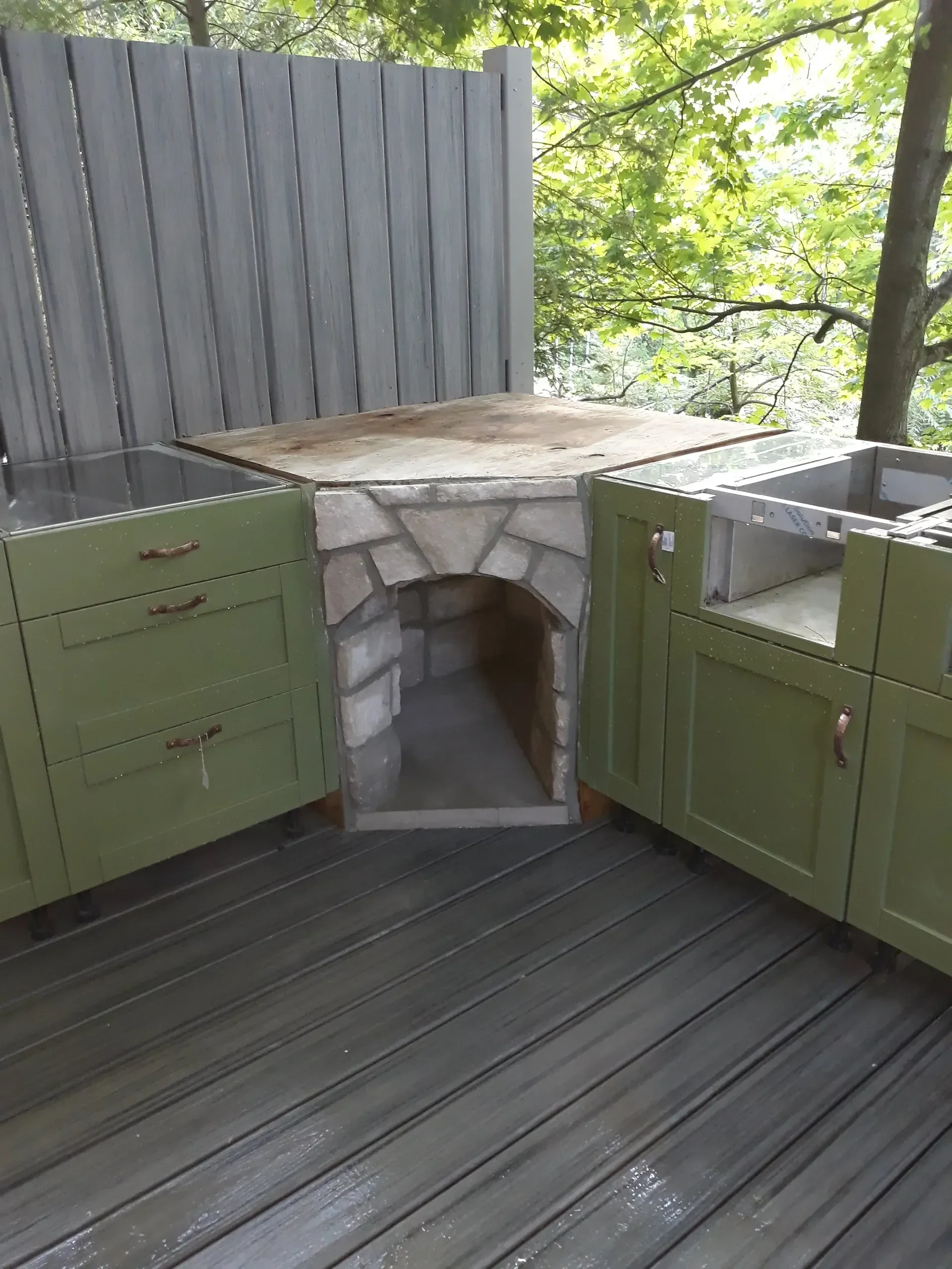An outdoor kitchen featuring green cabinets, a central stone masonry arch, and a temporary countertop is being installed on a dark composite deck surrounded by trees.