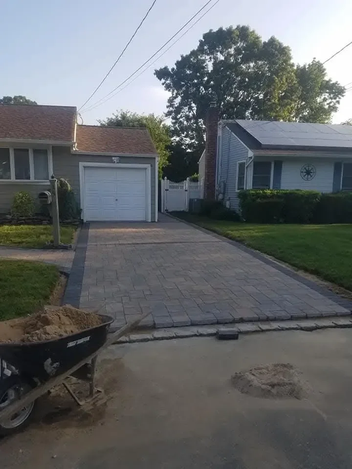 A newly installed paver driveway leads to a suburban garage, with a wheelbarrow full of sand indicating ongoing construction work in the foreground.