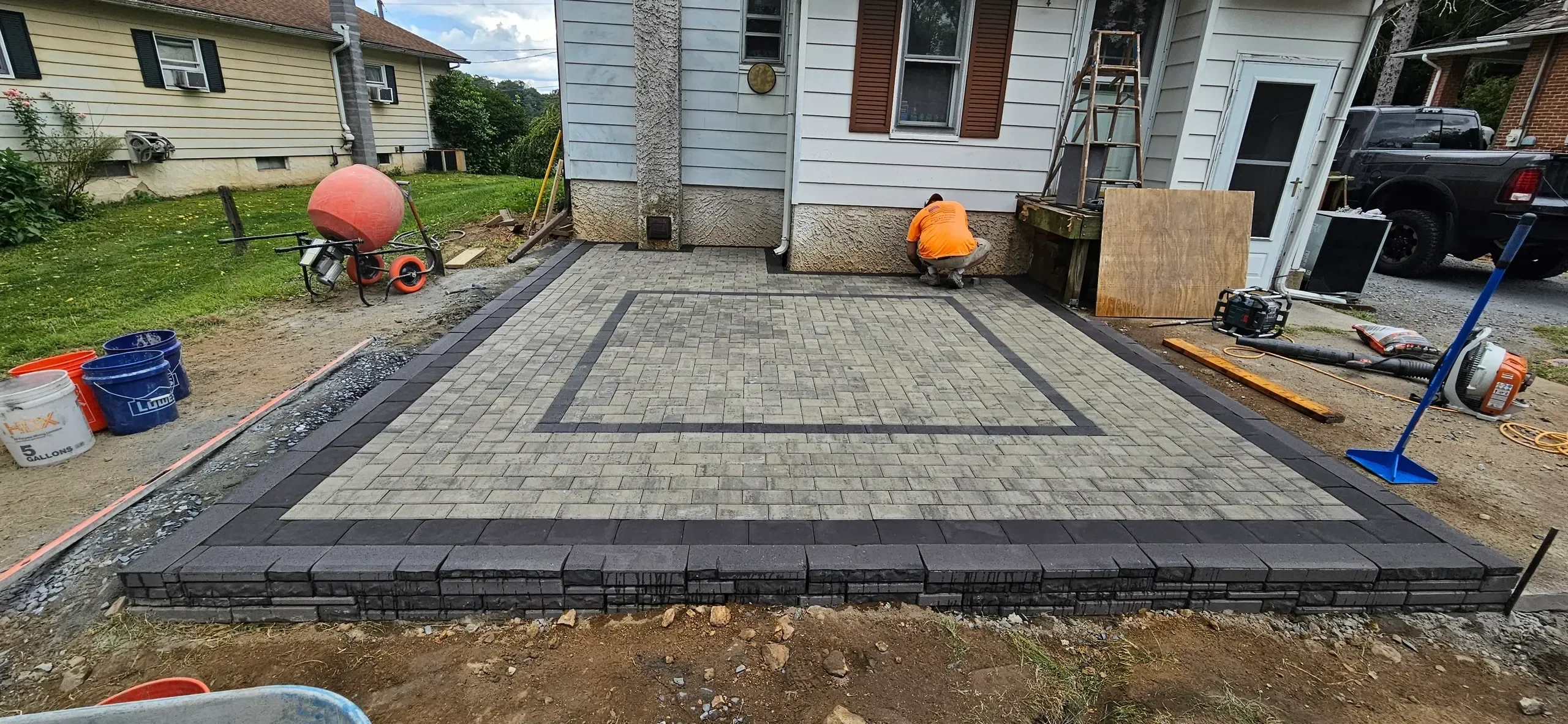 A worker installs the final edges of a newly constructed paver patio featuring light gray stones, a dark border, and a patterned inlay next to a residential home.