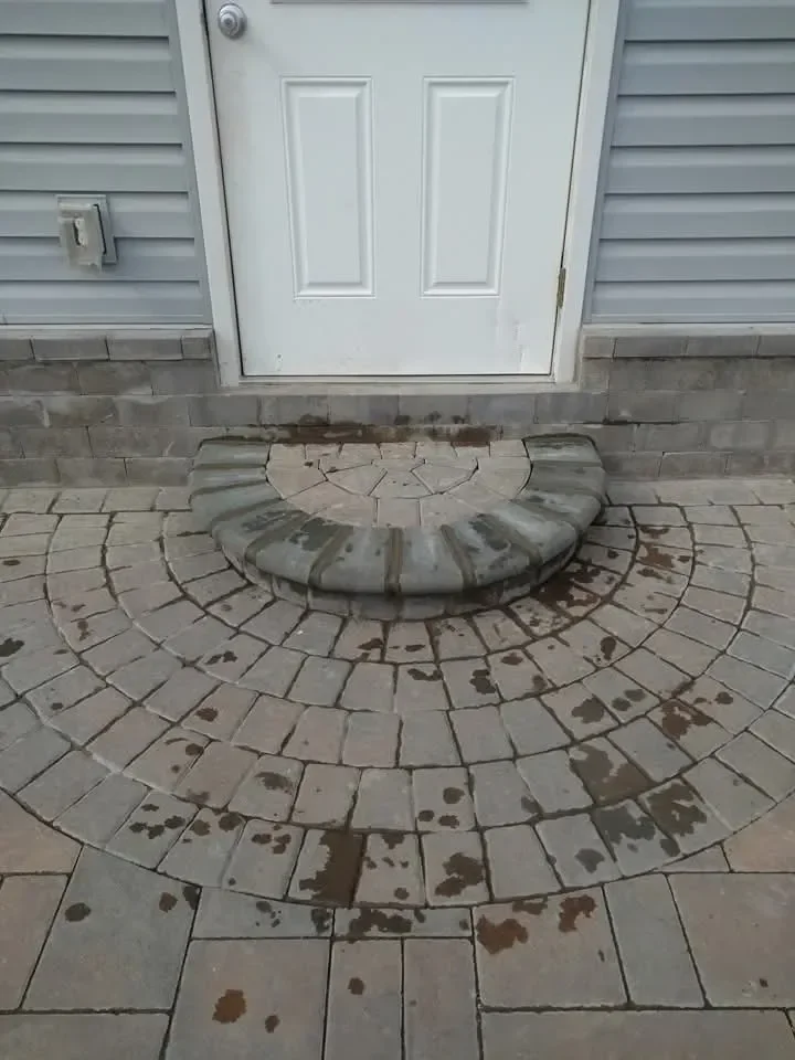 A newly installed paver patio featuring a radial pattern leads up to a curved stone step beneath a white exterior door and light gray siding.