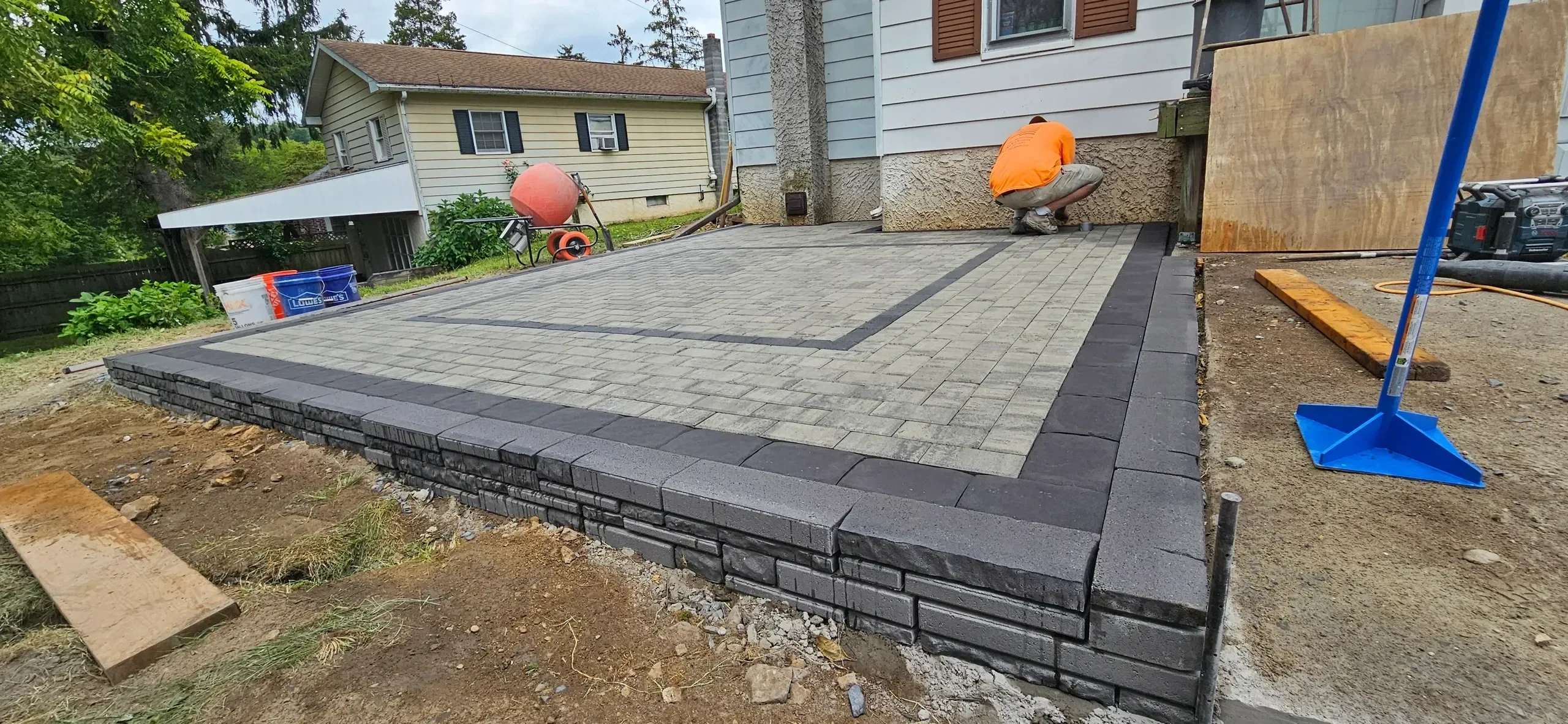 A worker installs the final details on a newly constructed raised paver patio featuring a dark gray retaining wall and contrasting paver colors.