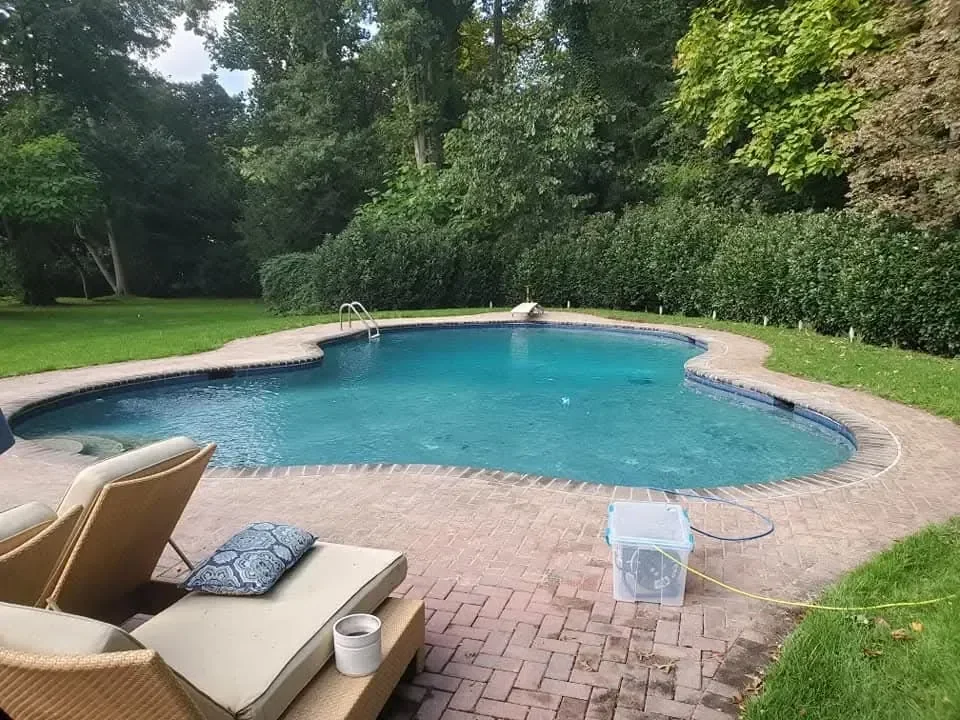 A curved residential swimming pool with turquoise water is bordered by a brick patio, lounge chairs, and lush green grass and trees.
