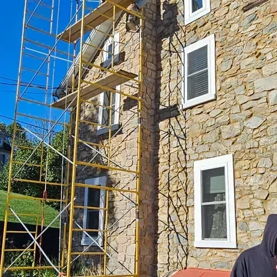 Yellow metal scaffolding stands against the rough stone facade of a building featuring multiple white-framed windows under a bright blue sky.