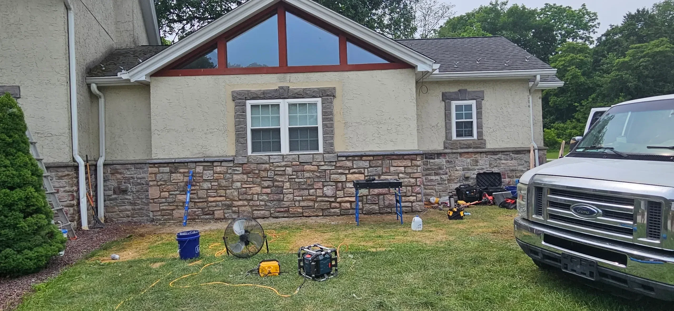 A residential exterior featuring stone veneer wainscoting and stucco walls is shown with construction tools and a work van on the lawn, indicating ongoing work.