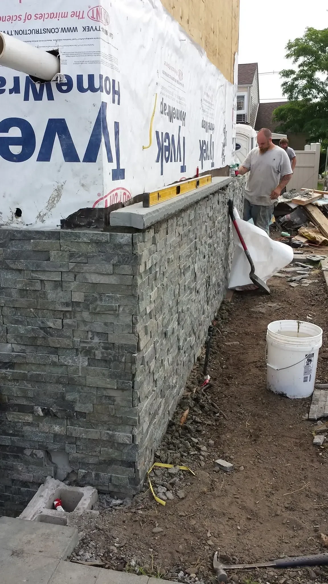 Workers install stacked stone veneer siding onto the lower exterior wall of a residential structure, utilizing a level and various construction tools.