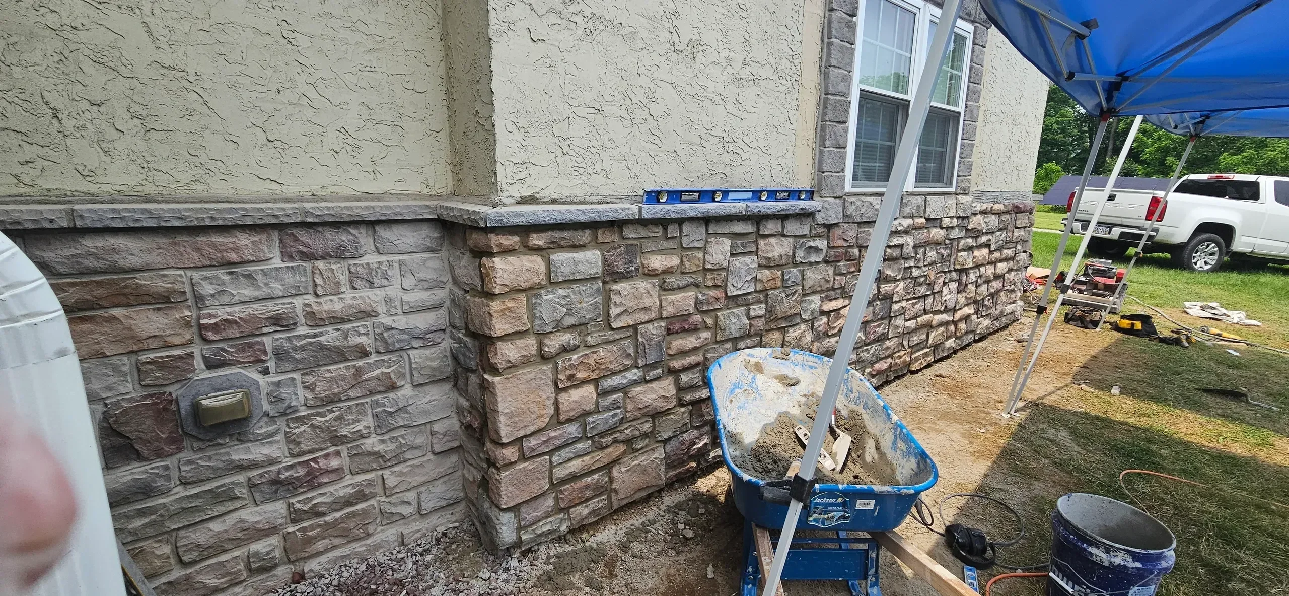 Installation of multicolored stone veneer siding is underway on the lower section of a textured residential exterior wall, with tools and materials visible nearby.