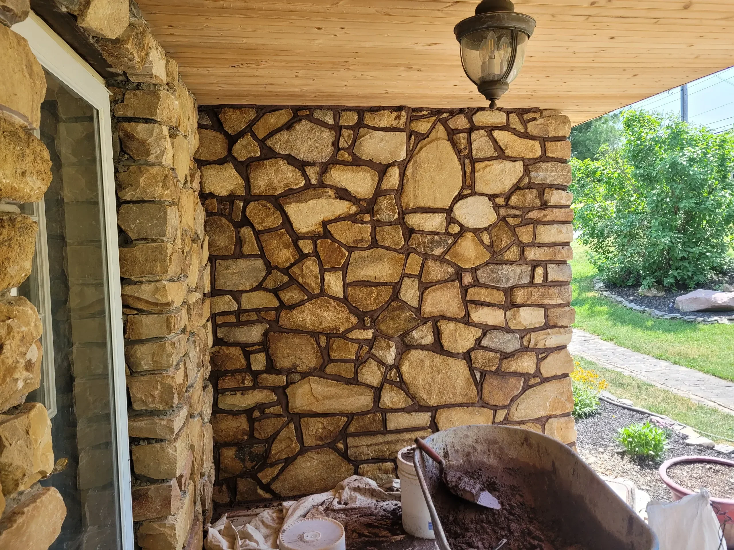 A close-up view of a stone veneer wall under a wooden porch ceiling shows construction materials, including a wheelbarrow filled with mortar, indicating ongoing masonry work near a window and outdoor landscaping.