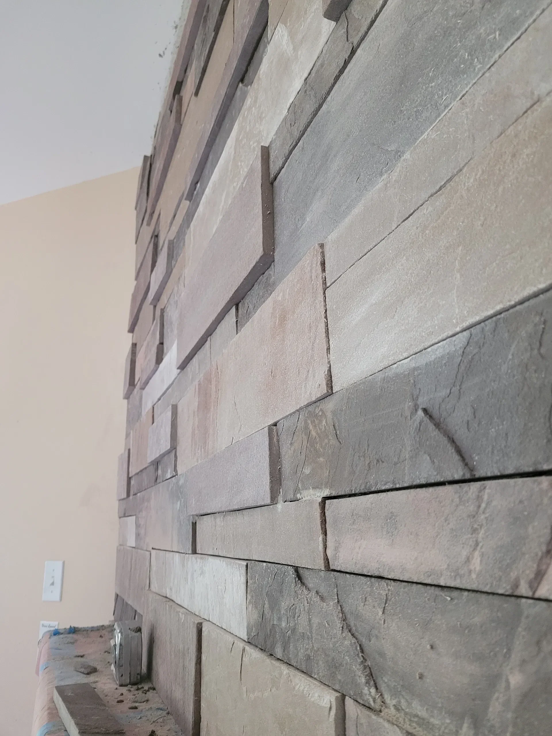 An angled close-up shot highlights the rough texture and varied earthy colors of stacked stone veneer being installed on an interior feature wall.