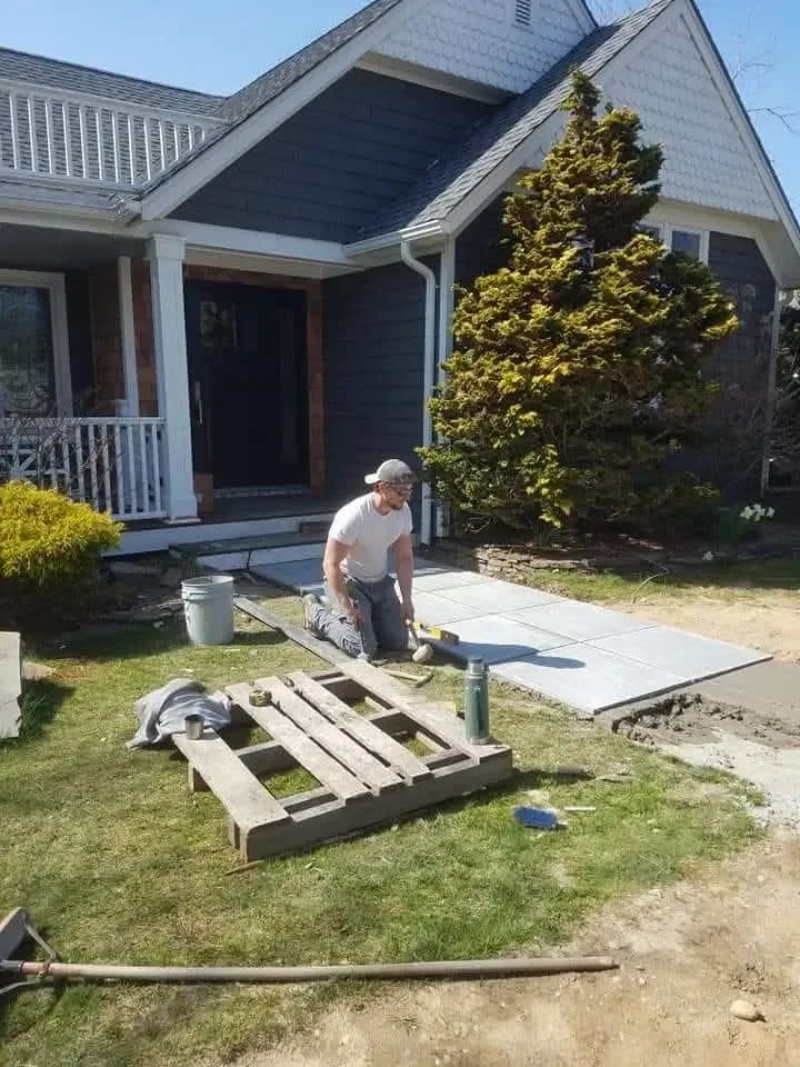 A construction worker kneels on the ground installing square pavers for a new walkway leading up to the front entrance of a residential home.